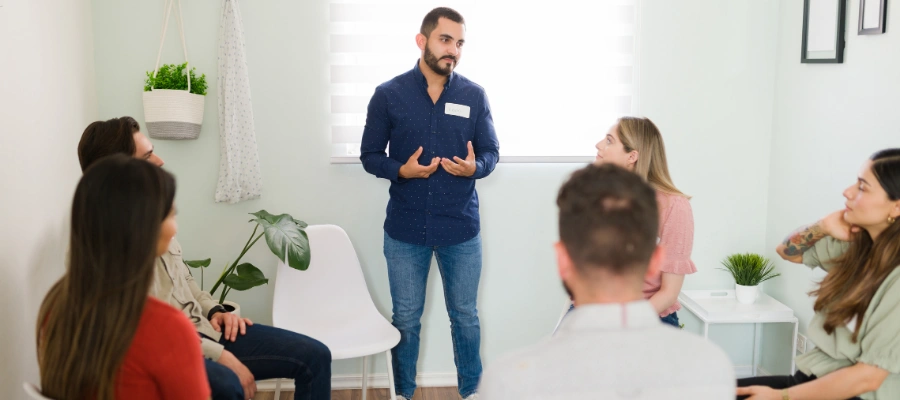 Man standing at an AA meeting with others seated looking on - What to Expect in First AA Meeting