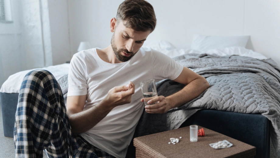 Image of a man sitting next to a bed holding a pill in one hand and a glass of water in the other - Can Antidepressants Help Addiction - Icarus Idaho