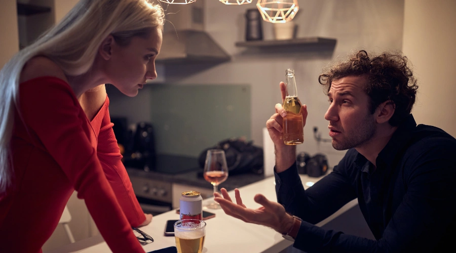 Image of a woman standing with her hands on a table, looking at her husband holding a bottle of beer and expressing a bewildered look on his face - Dealing With An Alcoholic Spouse - Icarus Idaho