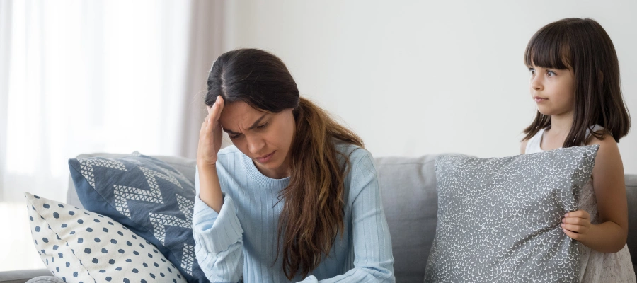 Image of an unhappy woman sitting on a sofa with her hand on her forehead, as her young daughter looks on, displaying  depression - signs of repressed childhood trauma in adults