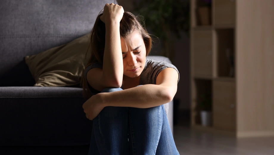 Image of a woman sitting on the floor with her eyes closed and knees up holding one hand on her head and displaying the Signs of Repressed Childhood Trauma in Adults - Icarus Idaho