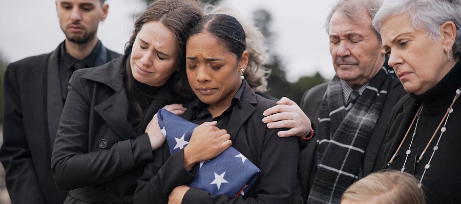 Image of a  Soldier’s wife dealing with grief and holding a flag at his funeral, being consoled by another female with men and a woman looking on