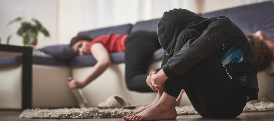 Image of man in a hoodie sitting on the floor with his knees next to his chest, beside a female sleeping on a sofa with an empty liquor bottle in her hand