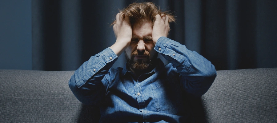 Image of a man in deep pain sitting on a sofa with both hands on his face - Withdrawal Symptoms of Inhalant Abuse