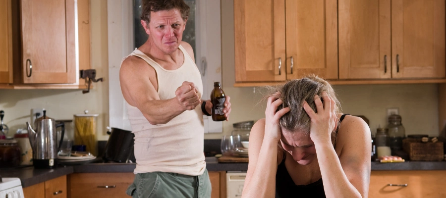 Image of a stressed partner sitting with both hands on their head as an unhappy man with a beer in one hand, clenching his fist, looks on, reflecting the emotional strain of being with someone struggling with alcoholism