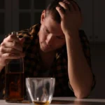 Image of a man sitting alone at a table in front of a bottle of whiskey and an empty glass - How Long Does Alcohol-Induced Depression Last - Icarus Wellness