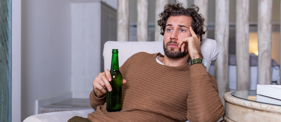 Image of a man sitting on a chair and holding a bottle of beer in one hand with a somber expression on his face