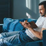 Image of a man sitting on a sofa, entering recovery passages into his journal - Recovery Journal Prompts - Icarus Wellness and Recovery