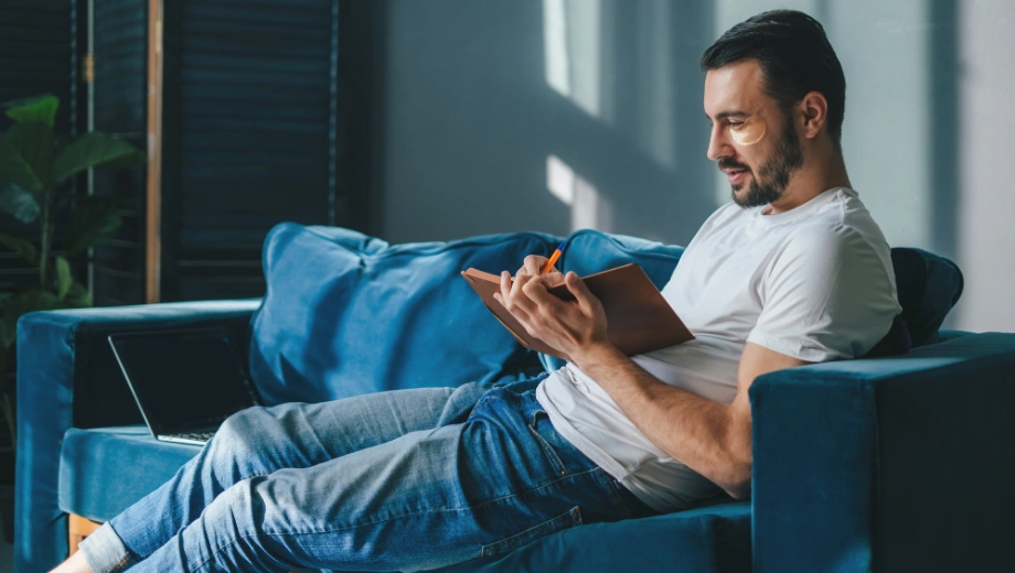 Image of a man sitting on a sofa, entering recovery passages into his journal - Recovery Journal Prompts - Icarus Wellness and Recovery