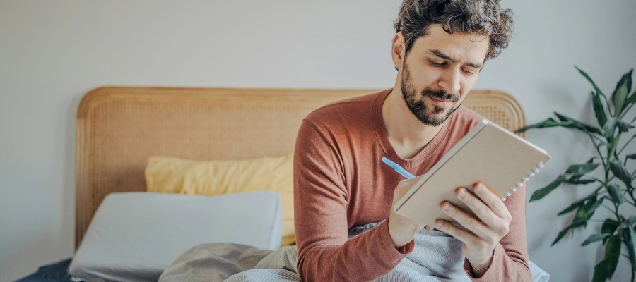 Image of a man sitting up in bed, journaling thoughtfully to build awareness of thoughts, emotions, and behaviors through REBT