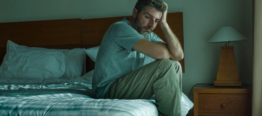 Image of a man in anguish, sitting on his bed and looking pensive, symbolizing the risks and challenges of exiting addiction treatment early
