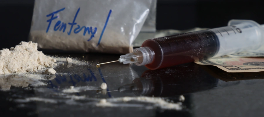 Image of a syringe on a table next to a bag labeled Fentanyl and a mound of white powder