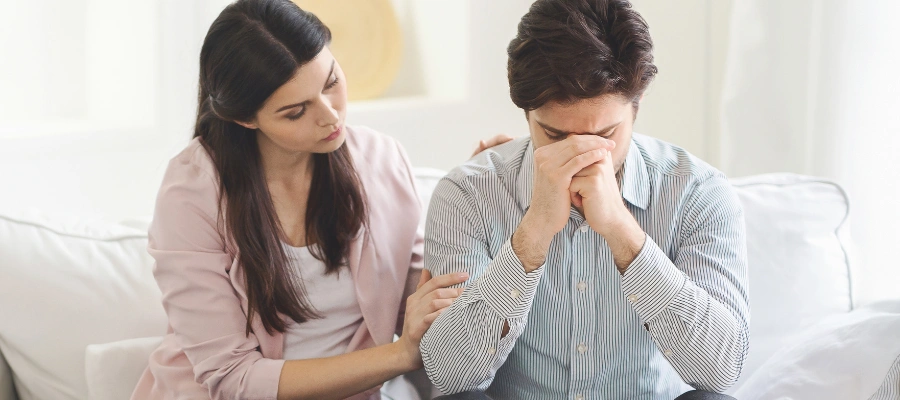 Image of a man with his hands on his face, appearing withdrawn and anxious while a supportive loved one looks on with concern